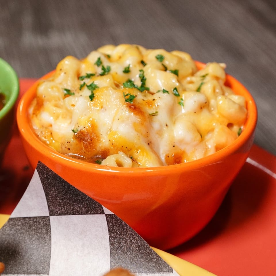 A vibrant orange bowl filled with creamy, golden-brown baked mac and cheese, garnished with fresh parsley.