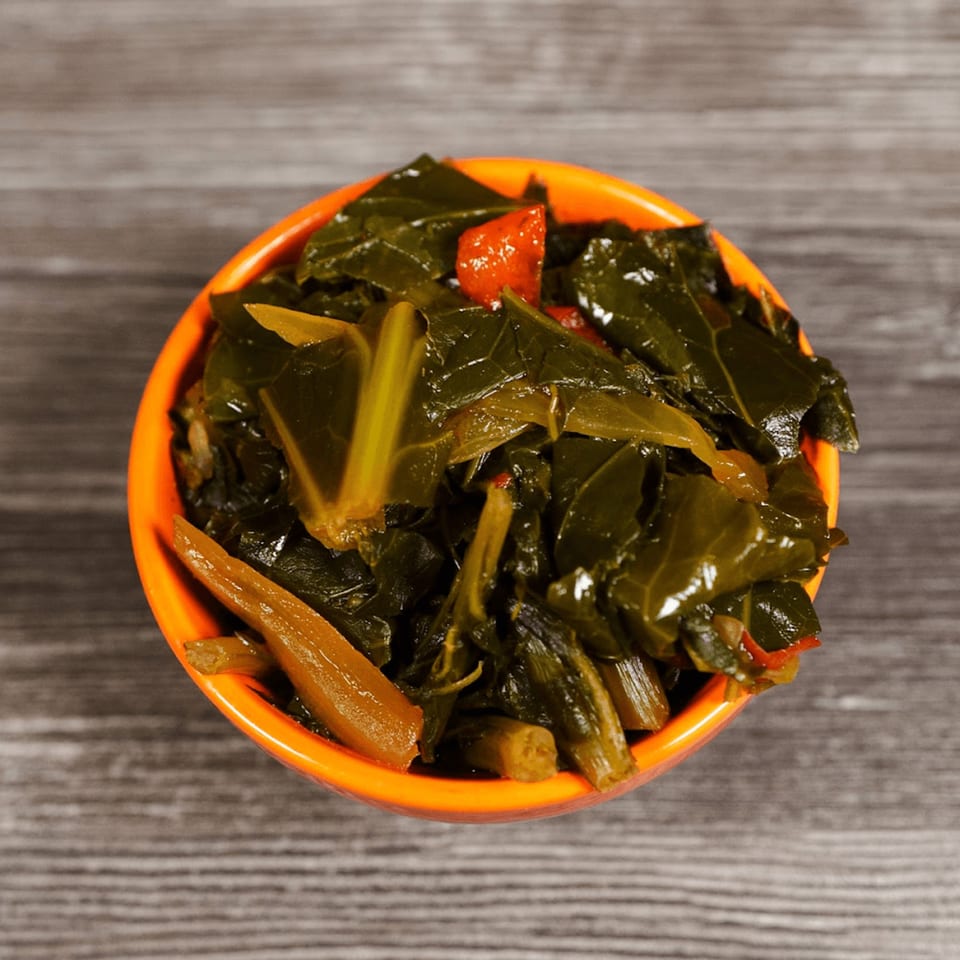 A vibrant orange bowl filled with cooked collard greens and red peppers on a wooden surface.