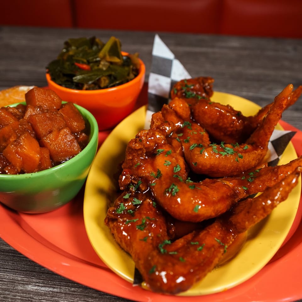 Soul food meal with glazed chicken wings, candied yams, and collard greens on a red tray.