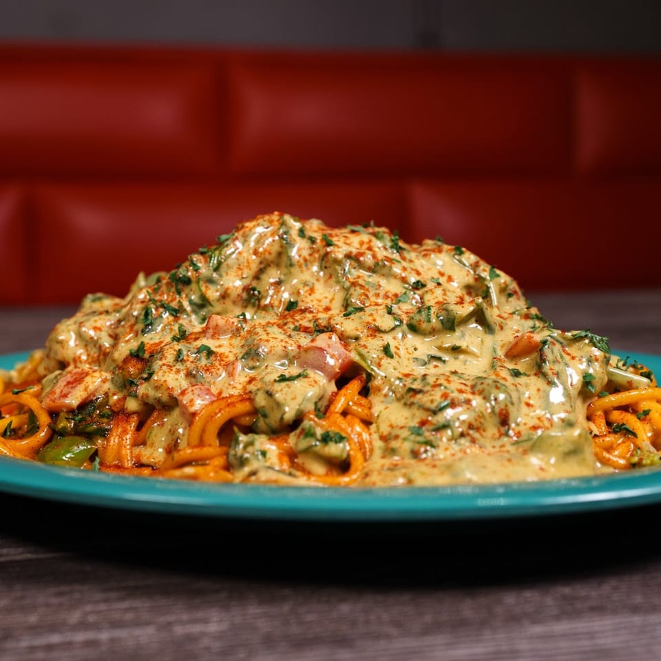 A vibrant soul-food dish of pasta with a creamy sauce, greens, and red spice on a teal plate.